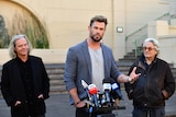 Chris Hemsworth stands at the microphone speaking, two other men stand either side of him