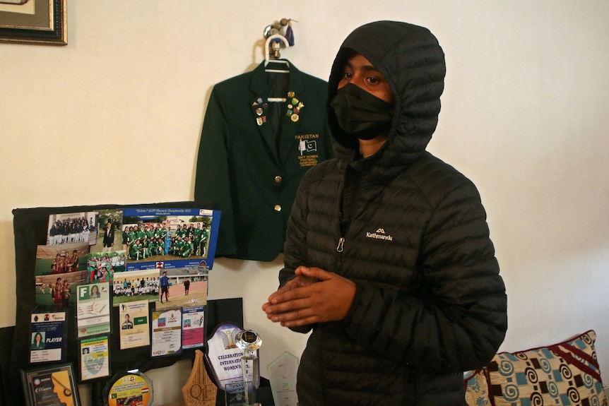 Symmya Kainat, in black mask and puffer jacket, standing in front of late friend Shahida Raza's sporting memorabilia.