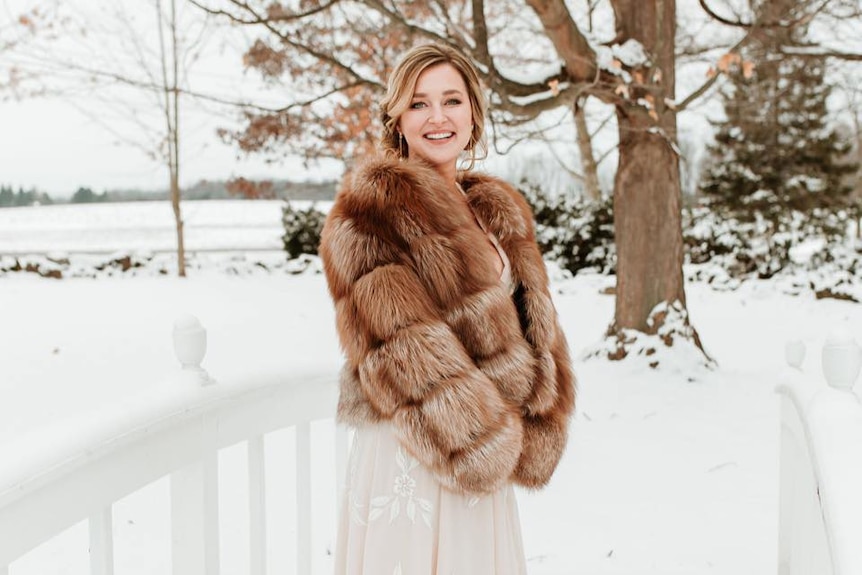 A blonde woman wears a brown fur coat over a white dress while smiling and standing on snowy field.