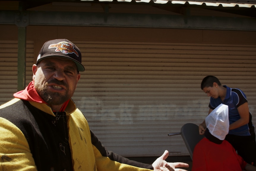 An Aboriginal man in hat and jacket stands facing the camera, a person adjusts a spit hood on someone in the background