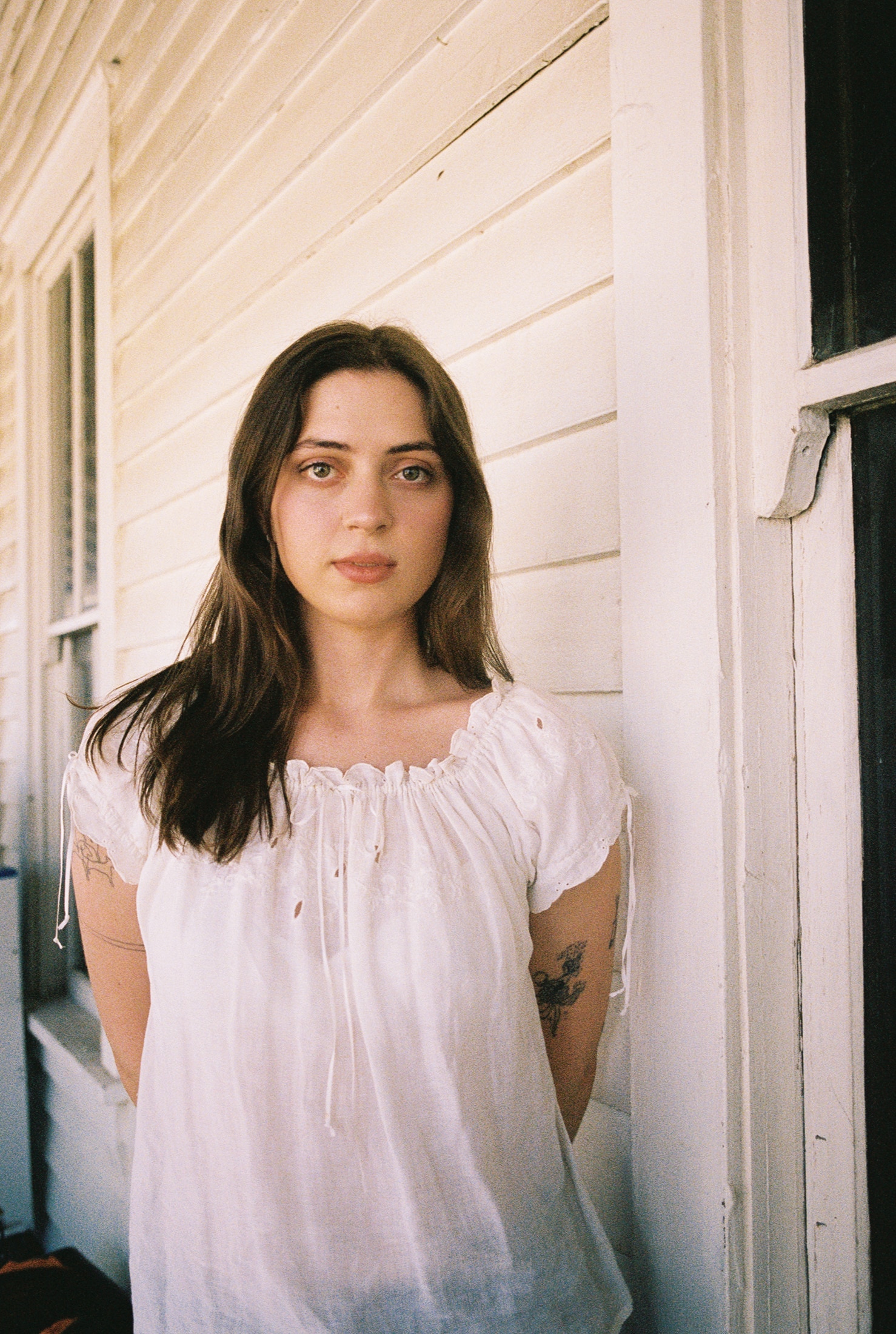 Lizzy McAlpine, with tattooed arms and long brown hair, stands on a weatherboard porch, looking at the camera.