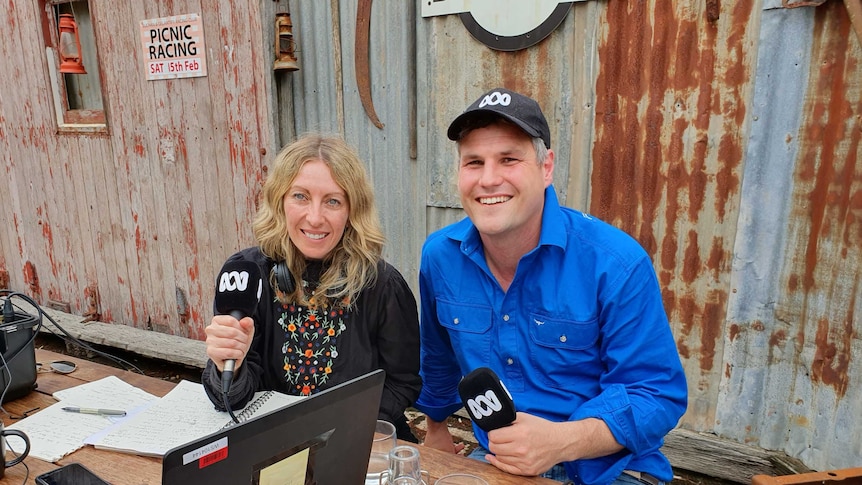 Richelle and Warwick sit at an outdoor table with steel shed walls behind them. They hold microphones, computers on the table.