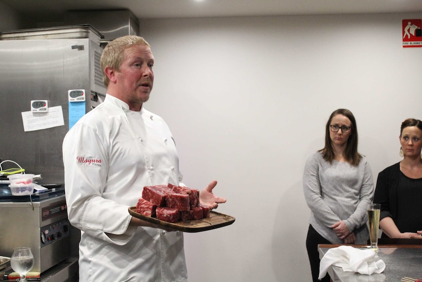 A man in a chefs outfit holding a plate of steak talking to guests