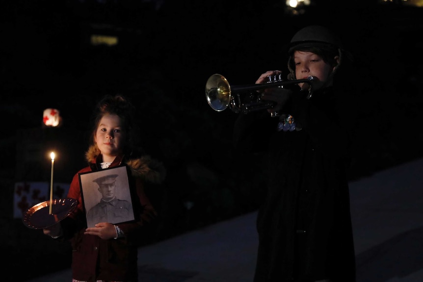 A boy plays a trumpet and a girl holds a candle and a photo of a serviceman as they stand in their driveway.