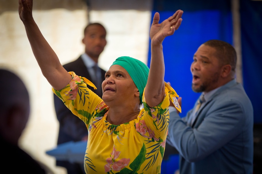 A woman in bright clothes and a headscarf dances with her hands in the air while others around her sing.
