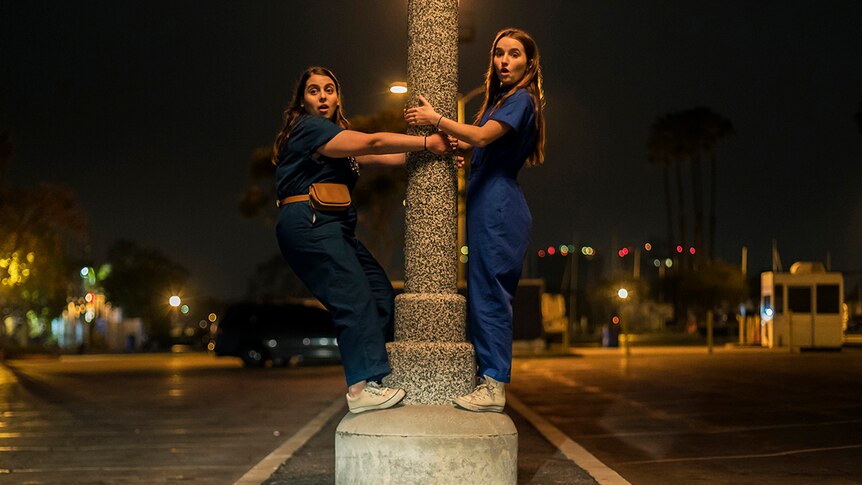 Colour still of Beanie Feldstein and Kaitlyn Dever clutching pole in the middle of the street at night in 2019 film Booksmart.