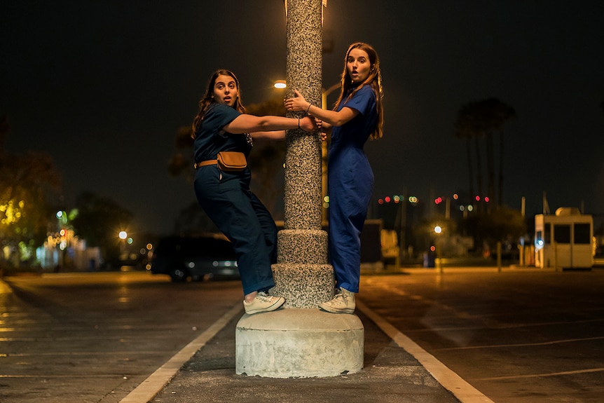 Colour still of Beanie Feldstein and Kaitlyn Dever clutching pole in the middle of the street at night in 2019 film Booksmart.
