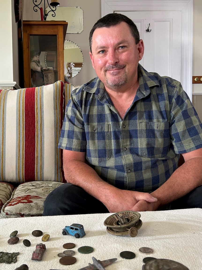 A man sits behind a table, smiling. On the table is a collected of rusted objects.