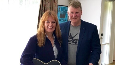 Jodi Ball and Dave Cox standing in the room of a house, with Jodi holding a black guitar.