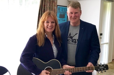 Jodi Ball and Dave Cox standing in the room of a house, with Jodi holding a black guitar.