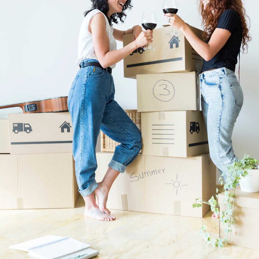 A female couple celebrate moving in together, they hold up two glasses of wine to say "cheers".