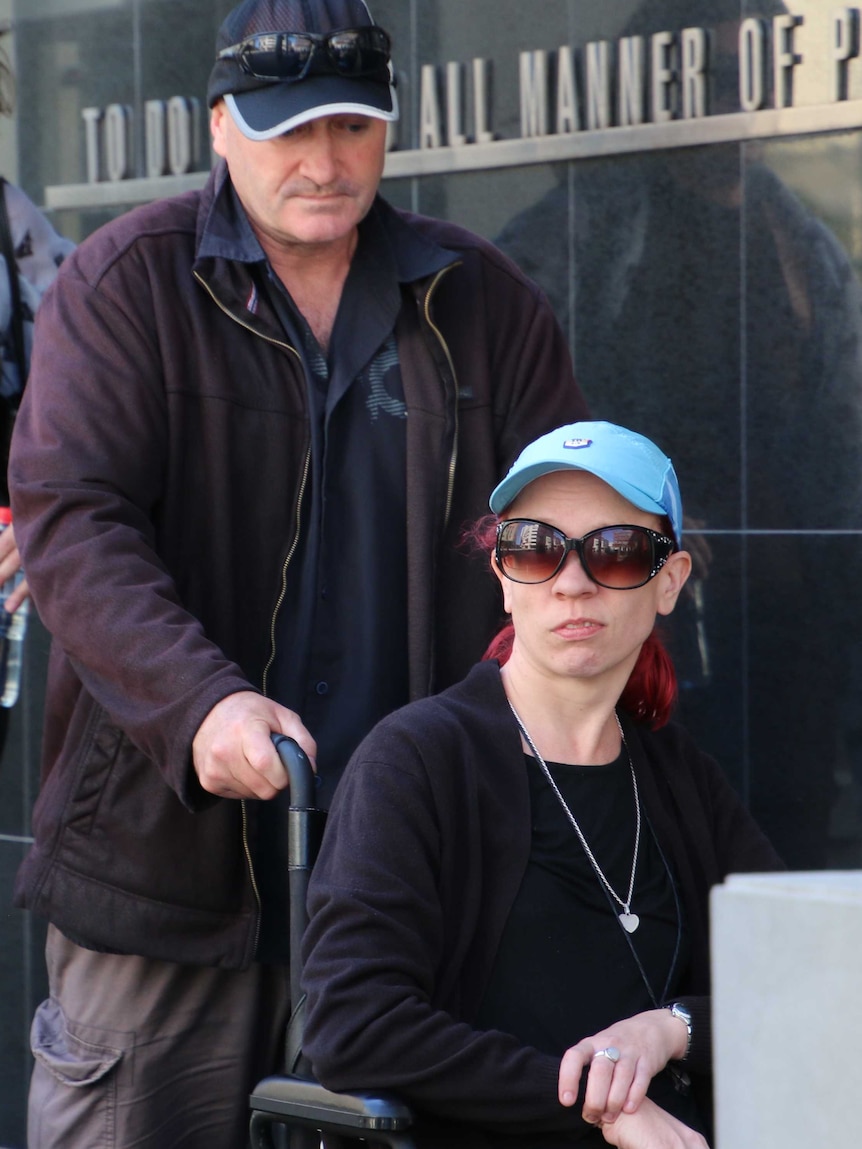 A man pushes a woman in a wheelchair down a ramp outside court.