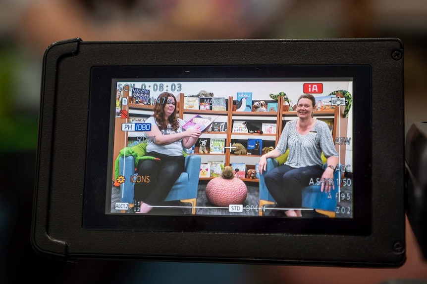 View of a video camera monitor showing two women in armchairs reading a children's book