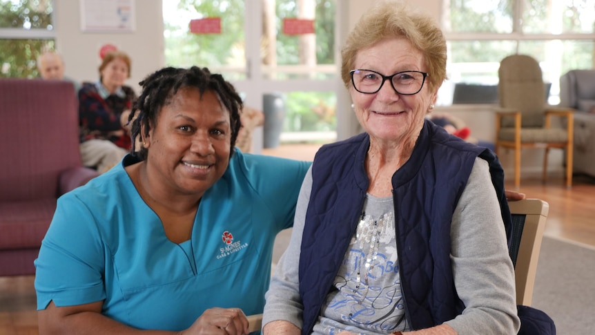 A Vanuatu woman in an aged care uniform crouched down next to an elderly resident, both smiling.
