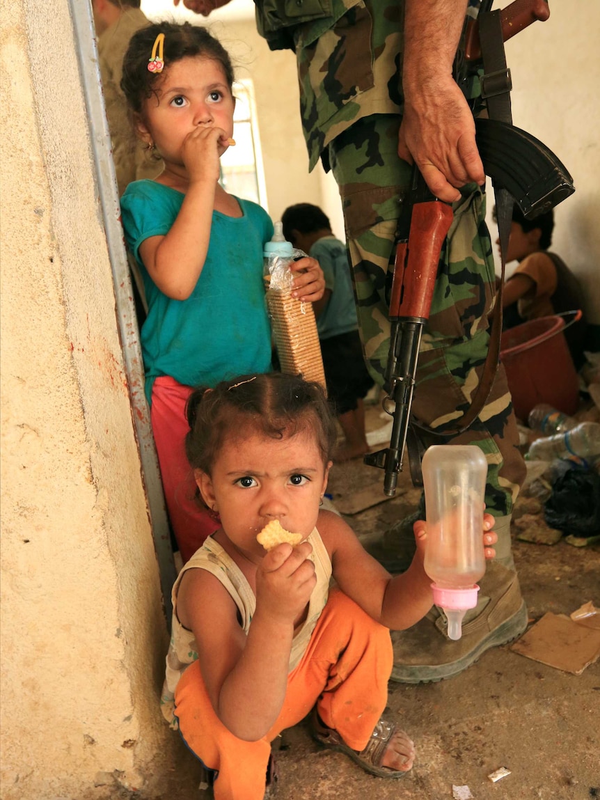 Two children holding bottles and biscuits stand in a doorway as a man holding a rifle walks behind them.