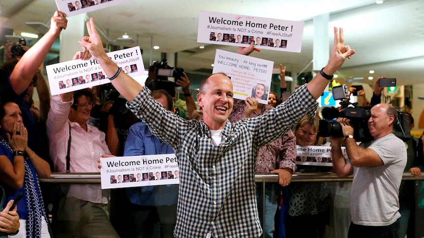 Peter Greste makes peace signs in front of a crowd of people holding signs saying "Welcome Home Peter".