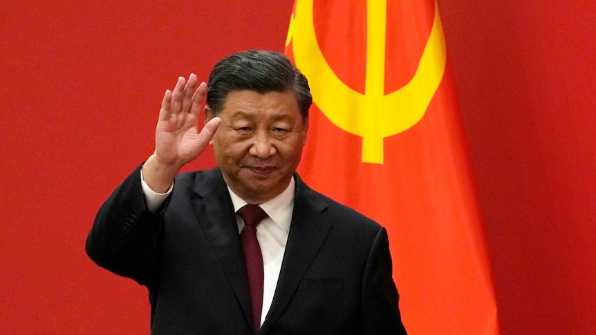 A suited man waves as he walks past a background of red drapes and a section of the Chinese flag.