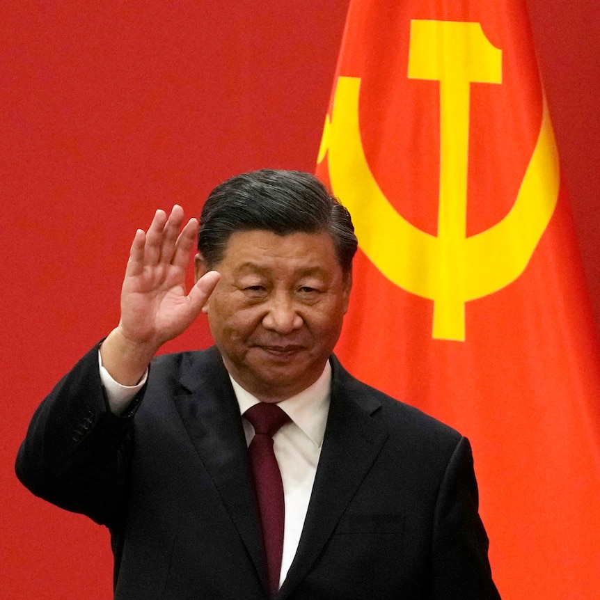 A suited man waves as he walks past a background of red drapes and a section of the Chinese flag.