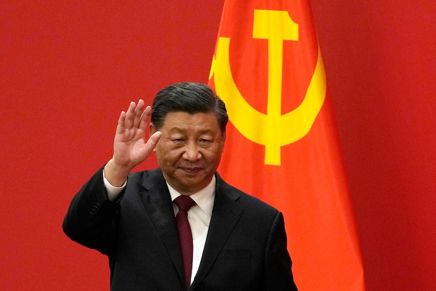 A suited man waves as he walks past a background of red drapes and a section of the Chinese flag.