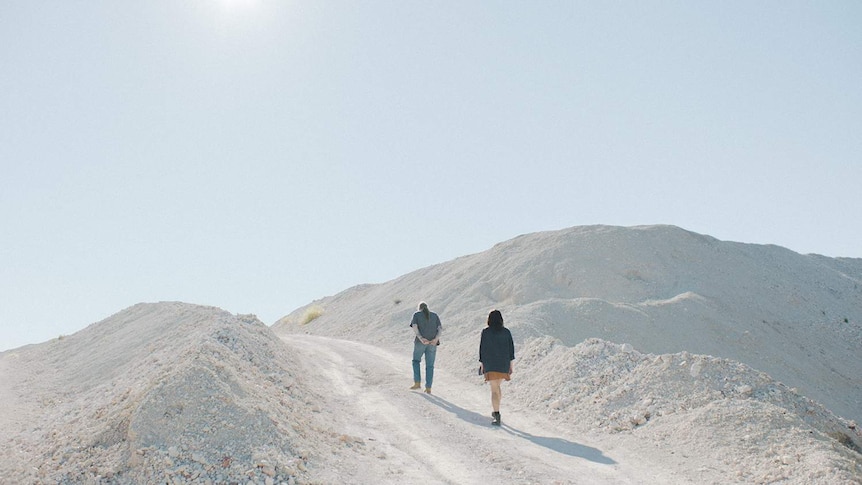 Two characters climb a stark, white pebble hill in the bright sunlight.