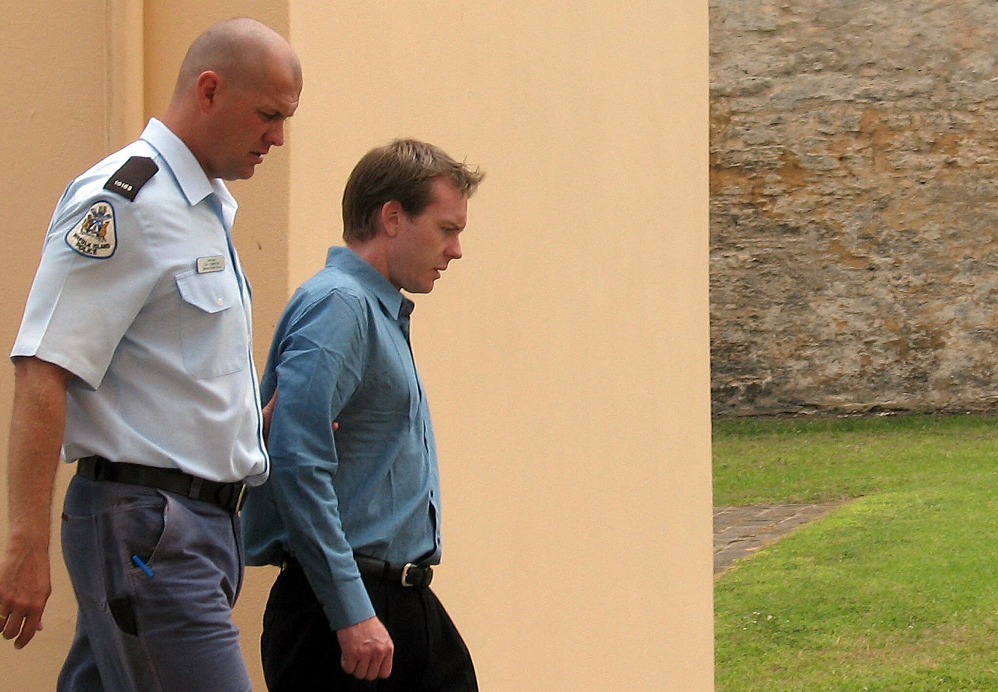 A police officer and a man in a blue business shirt walk in front of a concrete wall 
