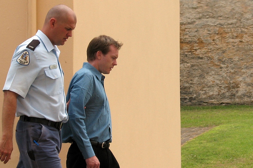 A police officer and a man in a blue business shirt walk in front of a concrete wall 