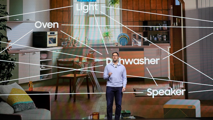 A man on stage against a screen showing a kitchen and living room