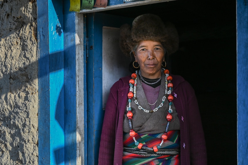 A woman stands in a blue-painted doorway wearing a fur hat, purple cardigan and strings of large beads