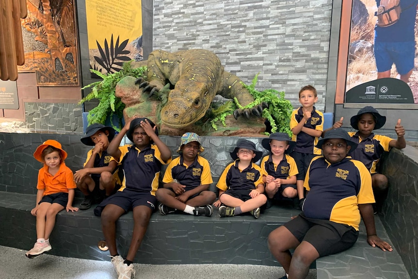 A group of school children sit on a rock, a dinosaur statue is behind them.