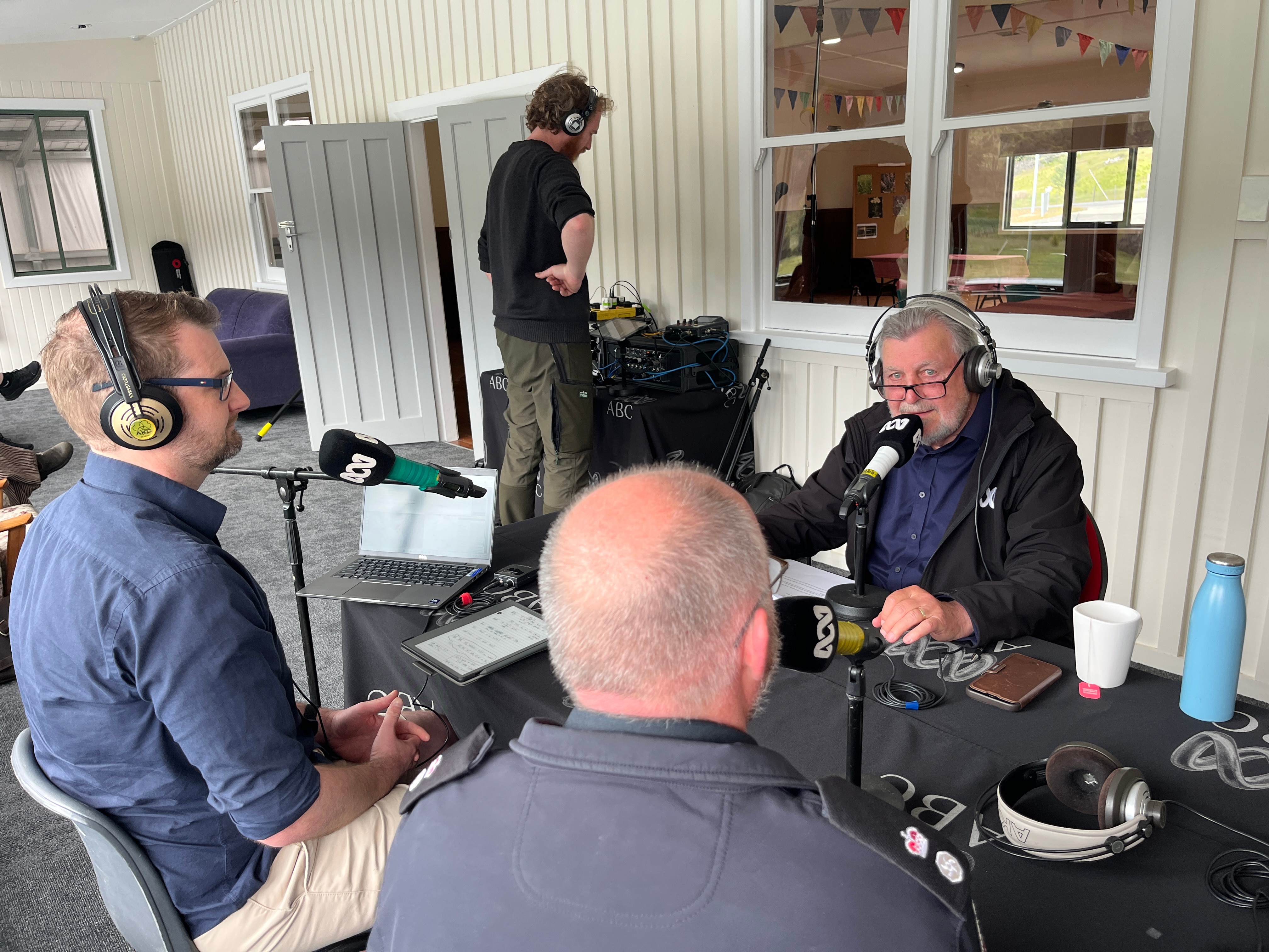 Man on microphone with two guests in amakeshift outside broadcats in a room 