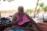 An Aboriginal woman sits on a sheet in scrub on the edge of Hedland