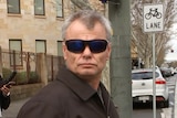 A man wearing sunglasses and a brown coat on a city street with a woman behind him