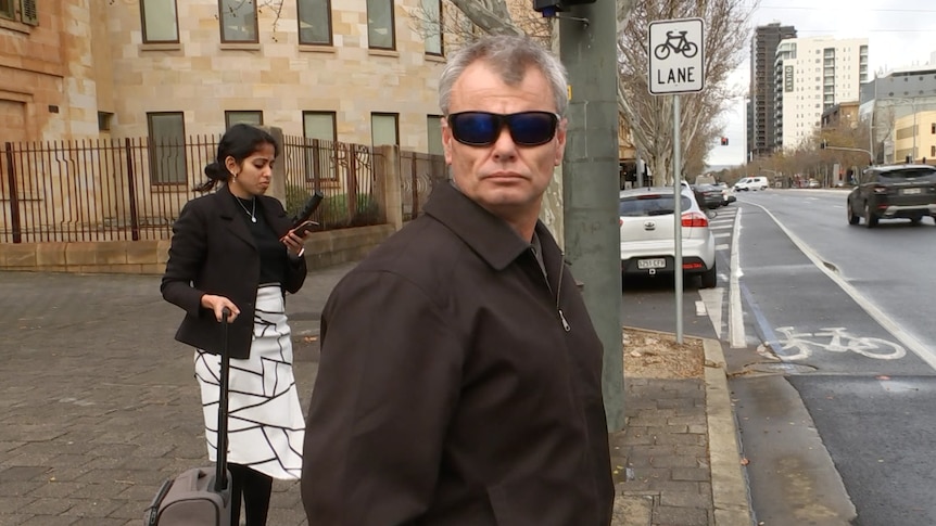 A man wearing sunglasses and a brown coat on a city street with a woman behind him