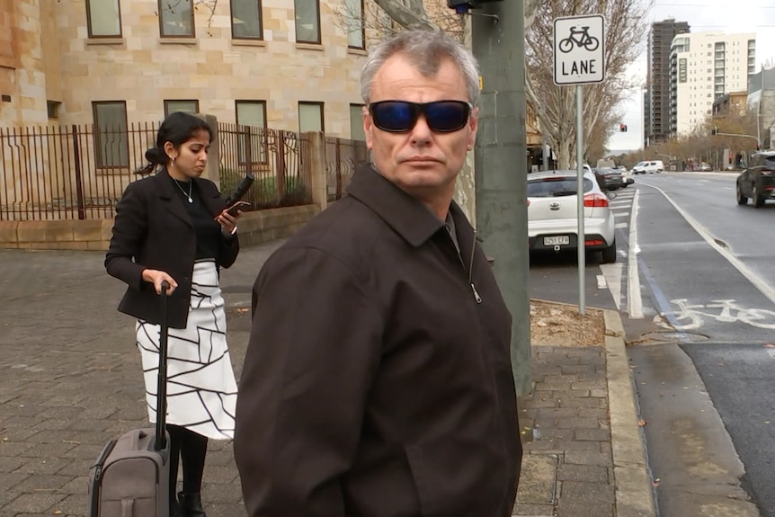 A man wearing sunglasses and a brown coat on a city street with a woman behind him