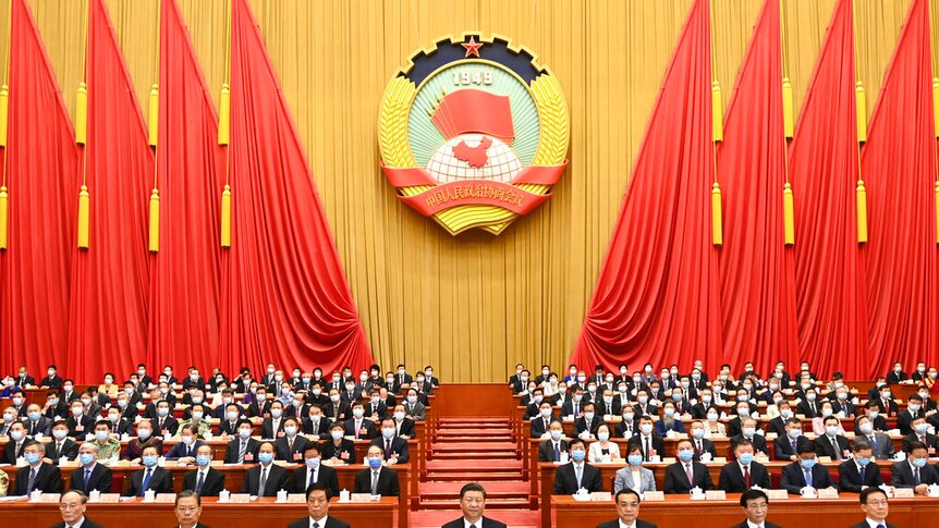 You view a wideshot of a grand red and yellow conference room with China's leaders seated in the foreground.