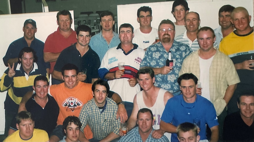 A group of 25 men in t-shirts and rugby jerseys smile at the camera.