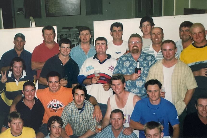 A group of 25 men in t-shirts and rugby jersey smile to the camera