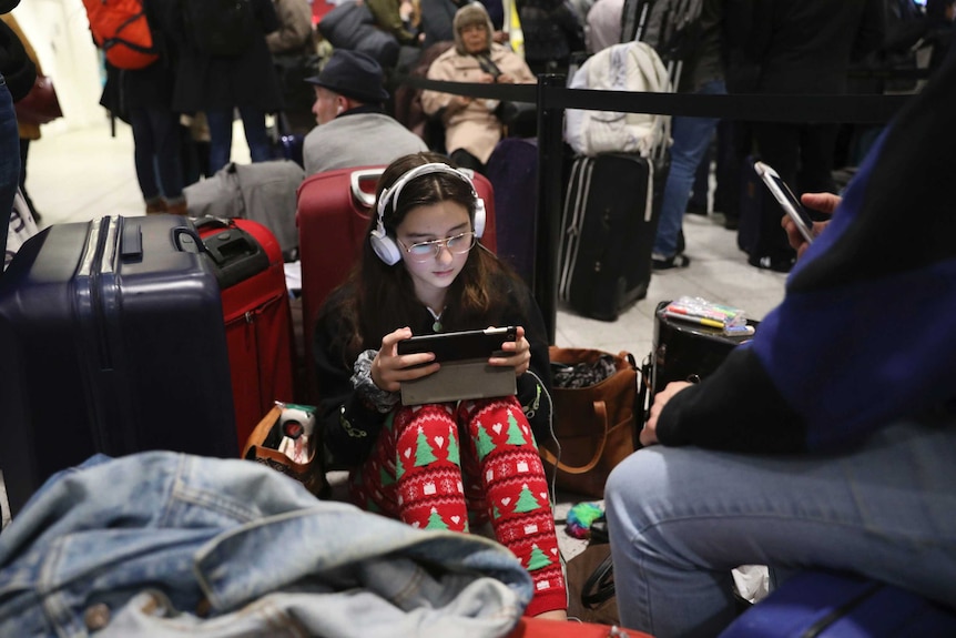A young girl sits on the ground in side Gatwick airport using an iPad with headphones on.