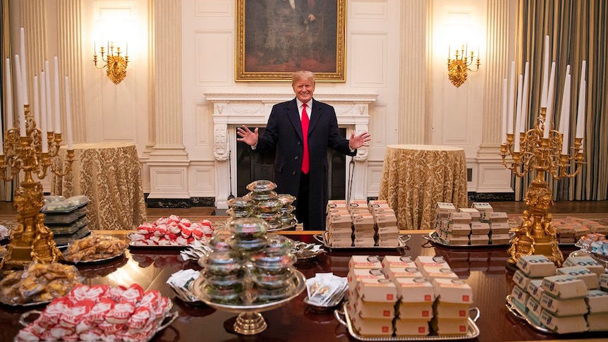 Donald Trump, with his hands out and a smile on his face, stands behind a table upon which sits massive piles of burgers.