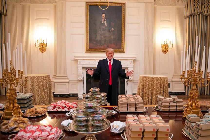 Donald Trump, with his hands out and a smile on his face, stands behind a table upon which sits massive piles of burgers.