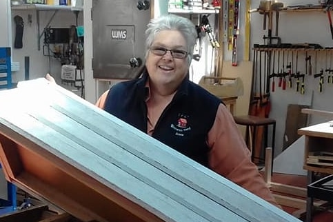 Middle aged woman with white hair in a shed holds up craftwork 