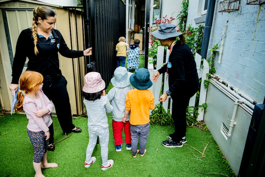 Two childcare workers are seen shepherding young kids through a gate 