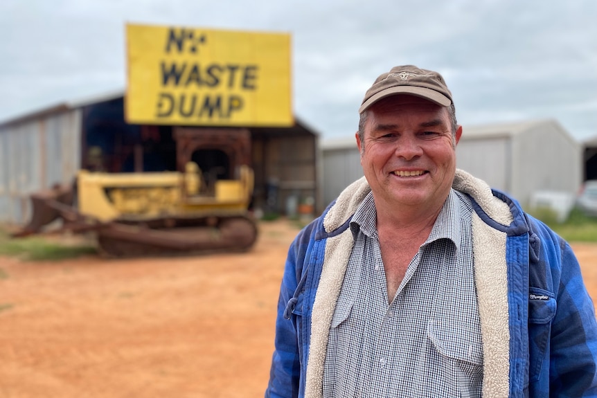 A smiling man in a baseball cap stands in front of a  shed with "no waste dump" on a sign.