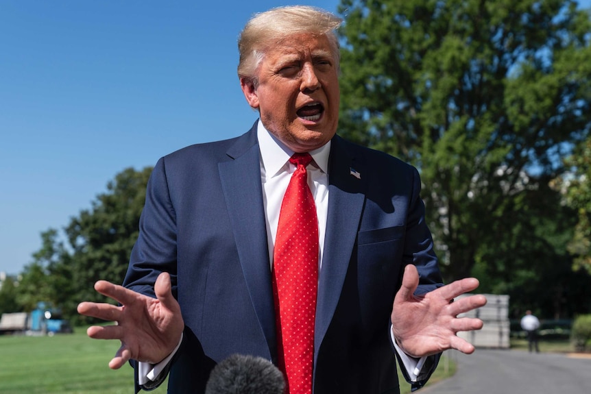 Trump dressed in a red tie holds his hands out as he speaks outdoors