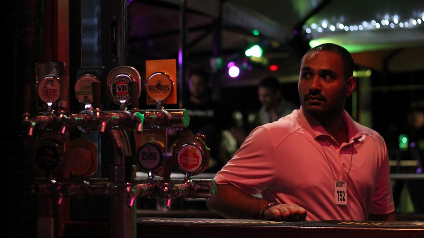 A young man leans on a bar with beer taps next to him at night.
