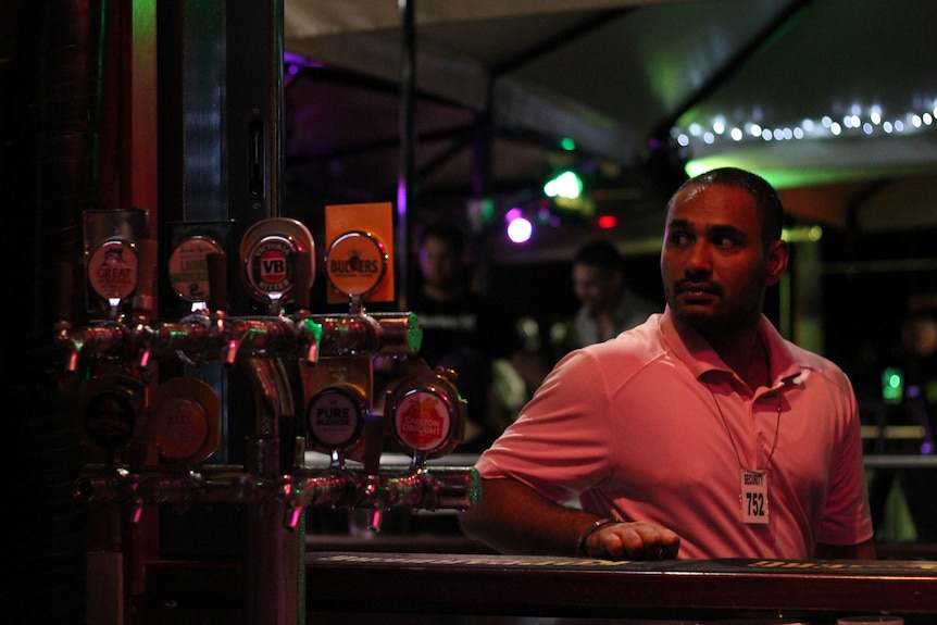 A young man leans on a bar with beer taps next to him at night.