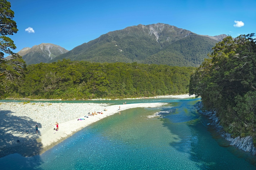 A blue lake in front of thick trees and a mountain. There are people lying on the sand next to the lake. 