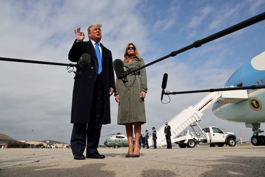 A man and woman in long coats, speak into microphones in the foreground, with Air Force One in the background.