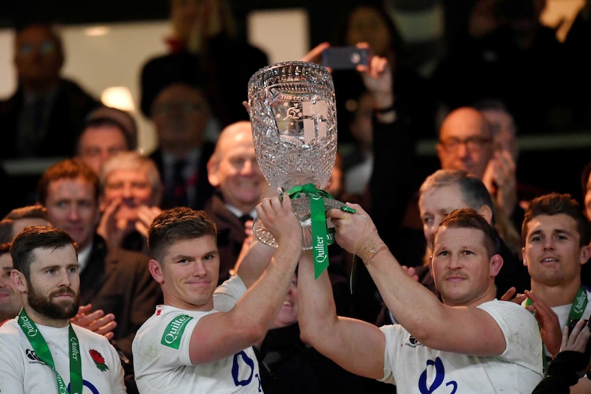 two men in white jumpers hold aloft a trophy with a crowd of people behind them