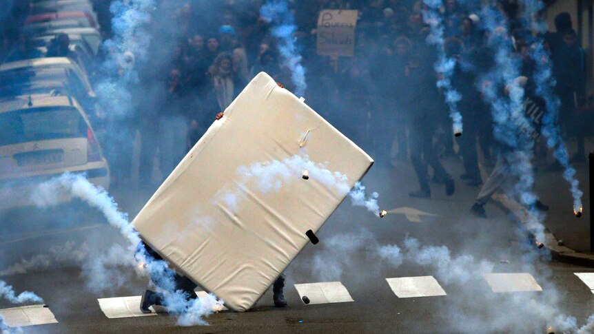 View down a street of youths taking cover behind a mattress from tear gas grenades.